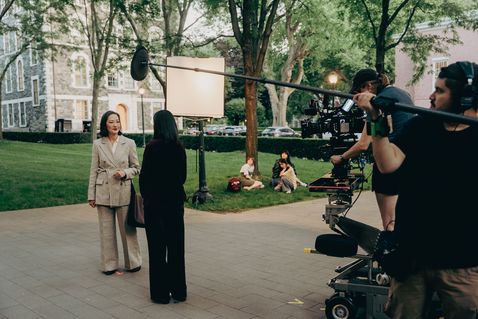Film crew staging an outdoor scene between two performers on a tree-lined campus walkway.