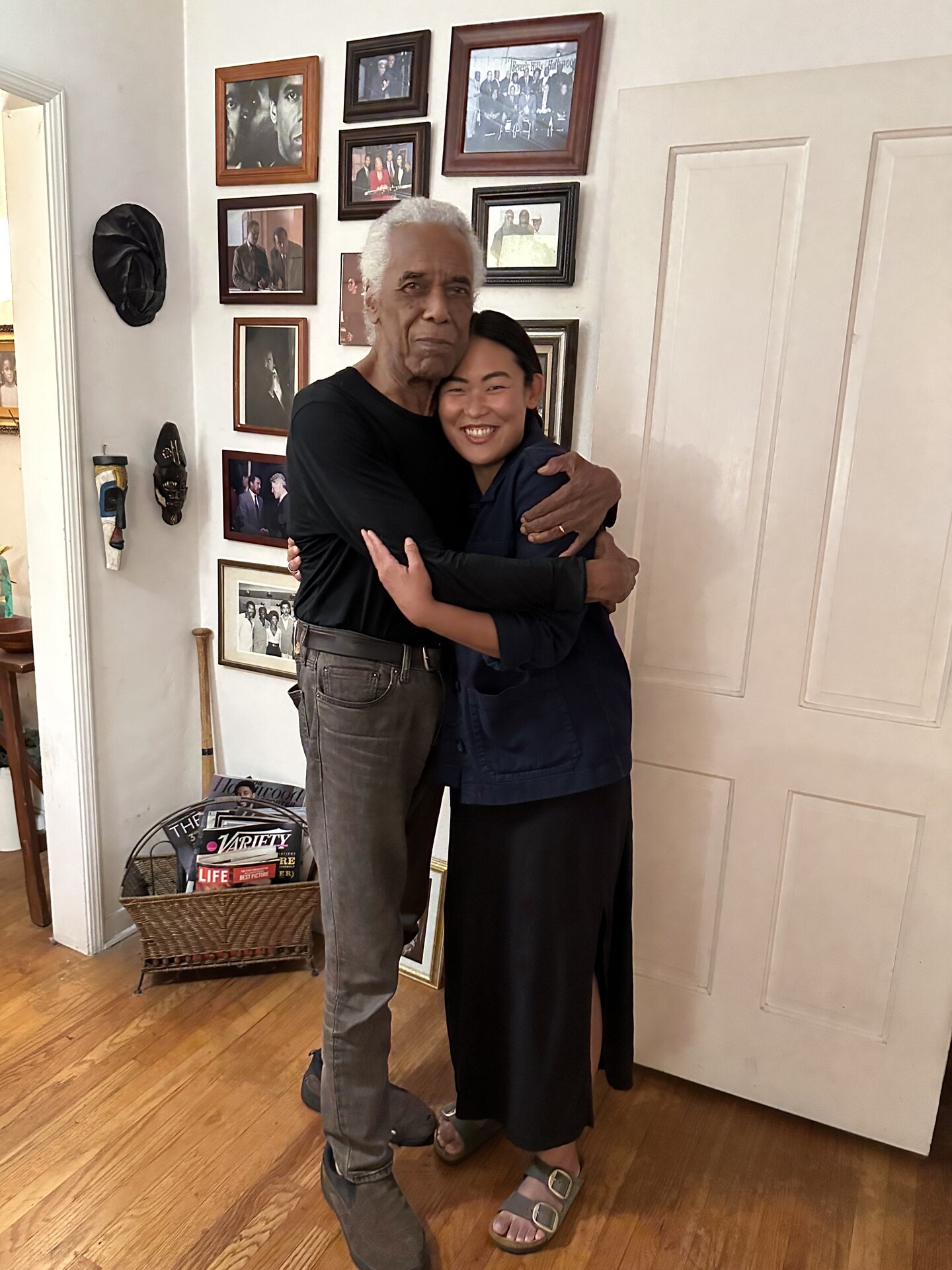 Robert Hooks and Grace Sun embracing inside a room lined with photographs.