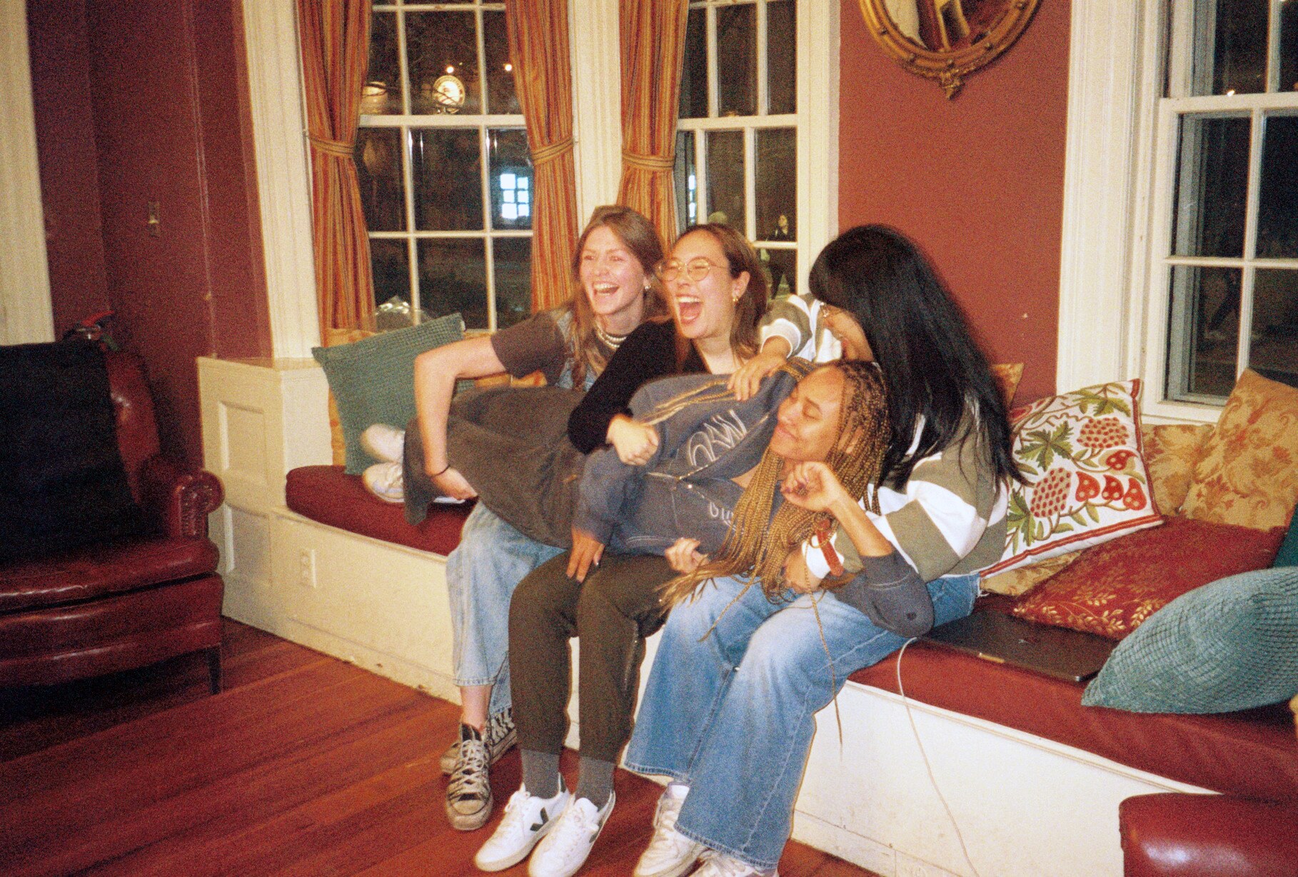 Four women laughing together on a window seat in a warm 35mm behind-the-scenes photograph.