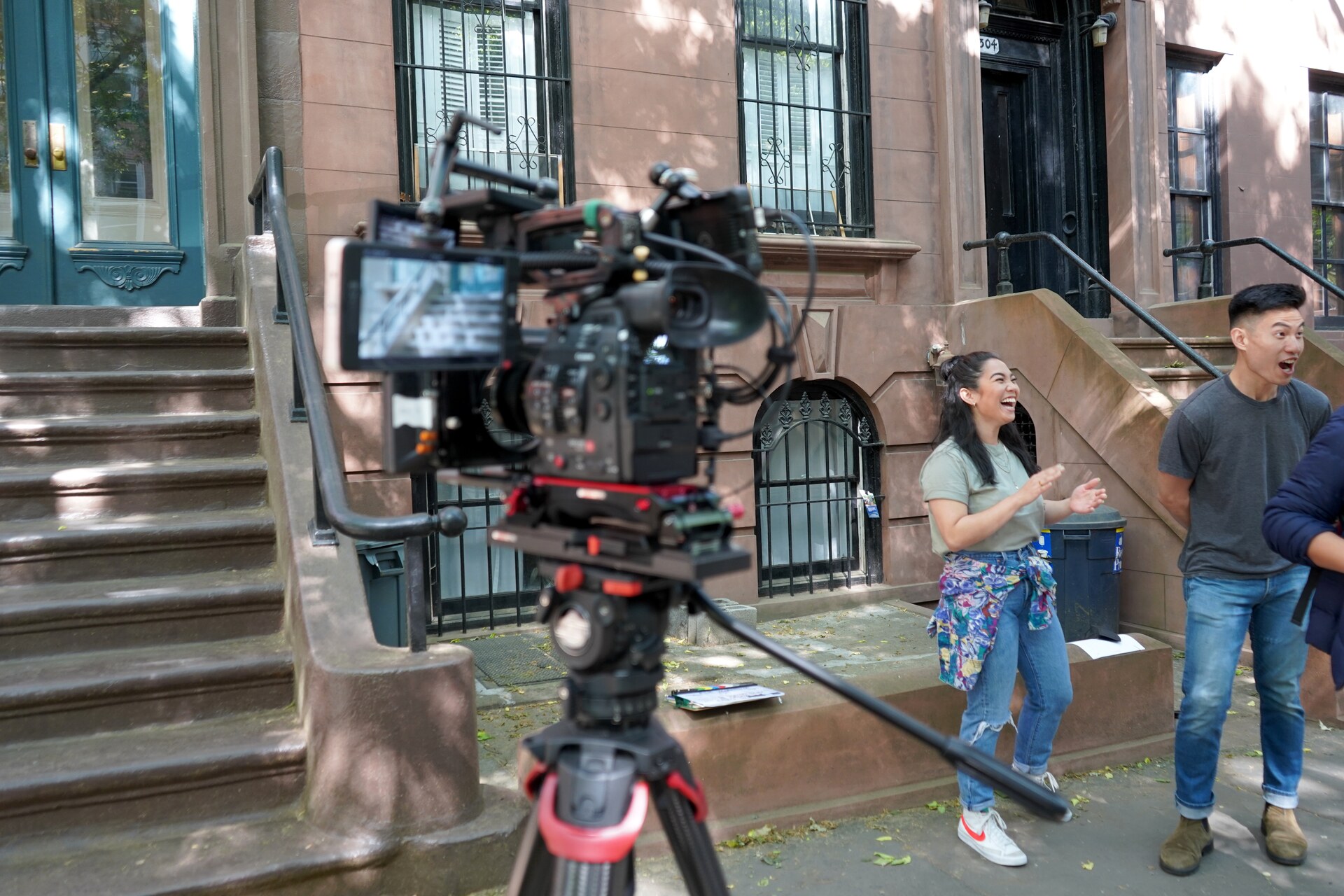 Two performers laughing on a Brooklyn sidewalk while a cinema camera records them.