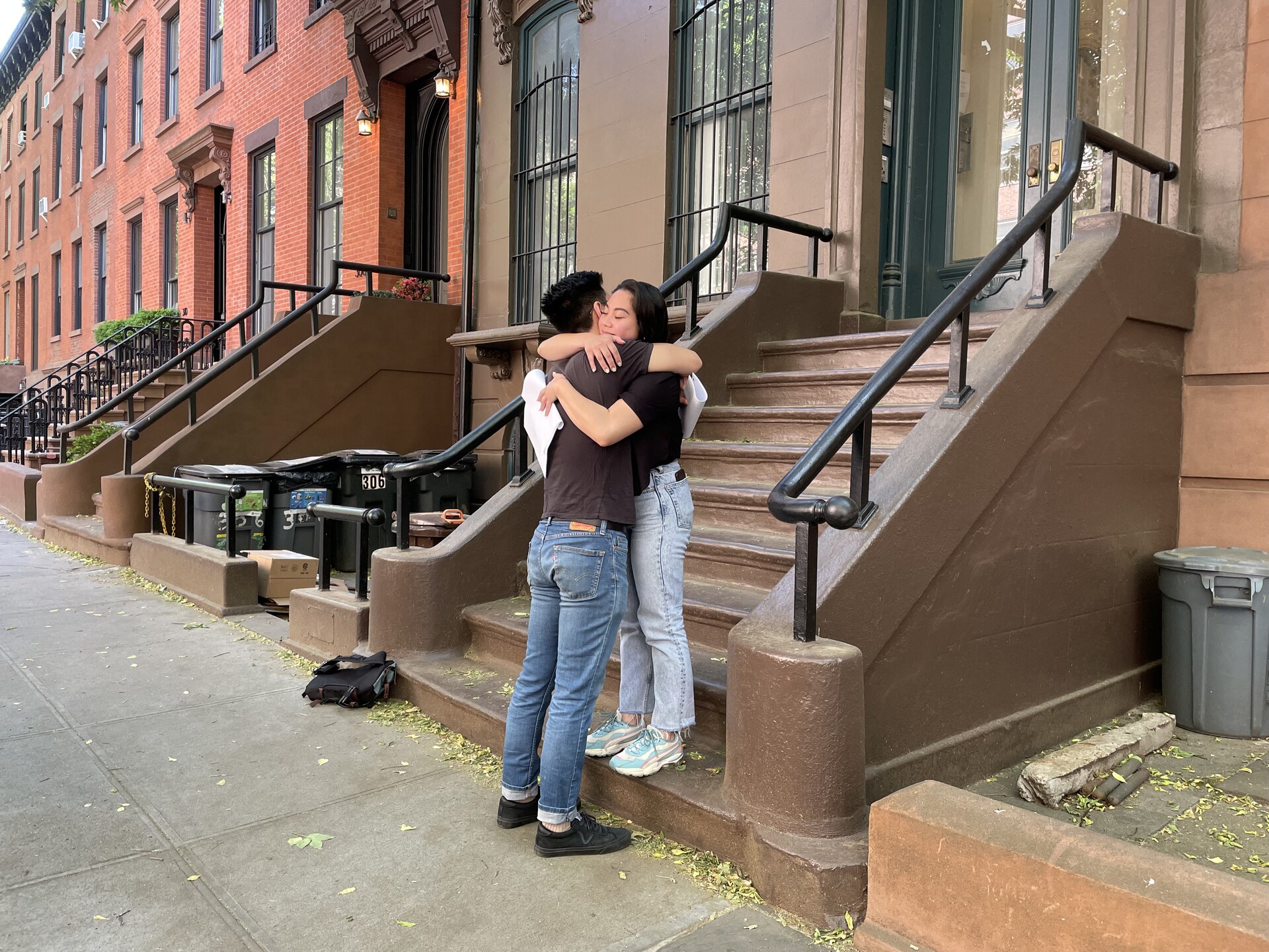 Two performers hugging on a brownstone stoop with production equipment nearby.