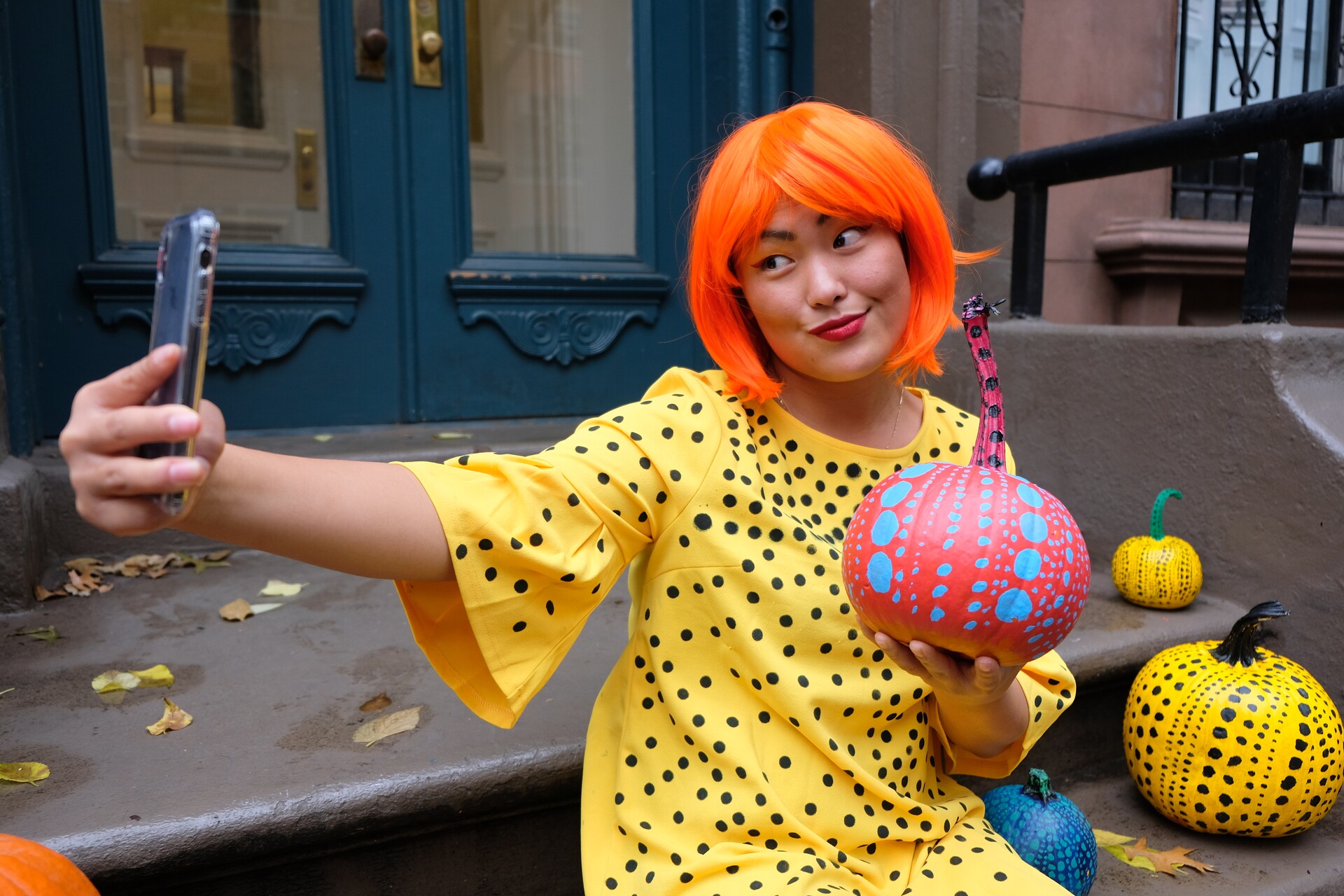 Grace taking a selfie in an orange wig and yellow costume while holding a painted pumpkin.